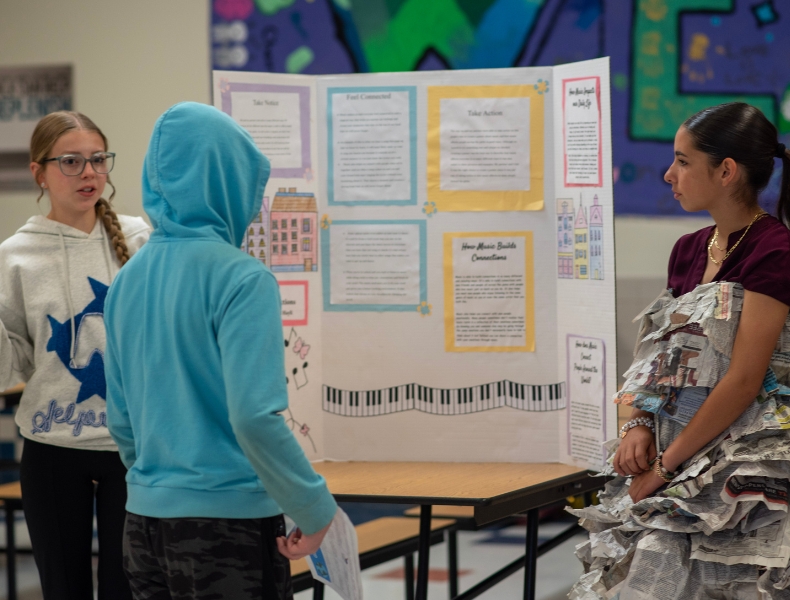 Three students standing in front of a trifold Global Leaders project poster. One student is presenting the poster.