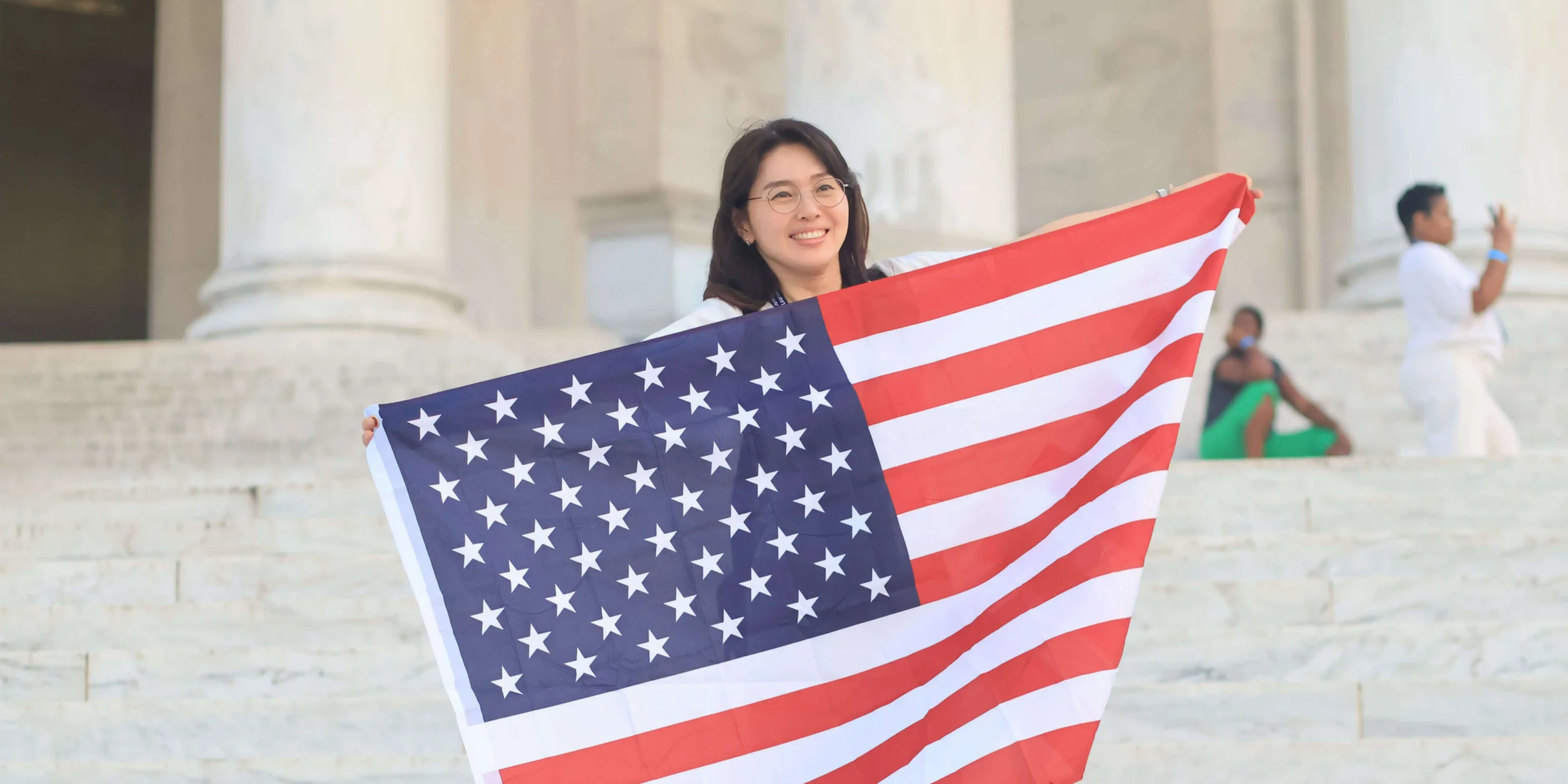 Smiling South Korean teacher holding an American flag in front of the Lincoln Memorial in Washington, D.C., showcasing her arrival in the USA through the J-1 visa cultural exchange program to teach in American schools.