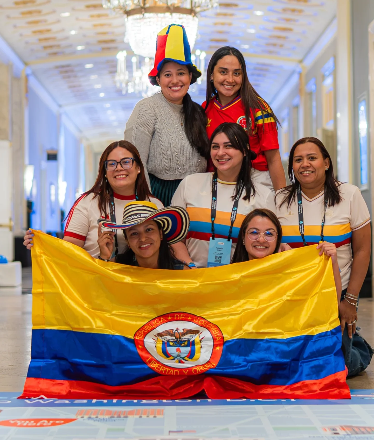 A group of teachers from Colombia pose joyfully in a hallway holding a large national flag, celebrating cultural exchange and their opportunity to teach in the U.S. The image highlights unity, diversity, and professional growth through teaching abroad.