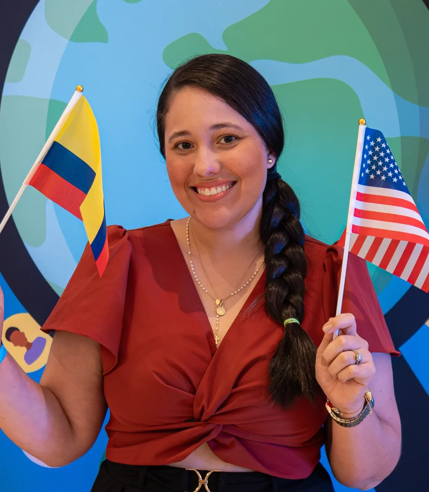 Female teacher smiles while holding both the Colombian and U.S. flags in front of a globe-themed backdrop. The photo reflects cross-cultural exchange, teaching in the United States, and professional development for educators abroad.