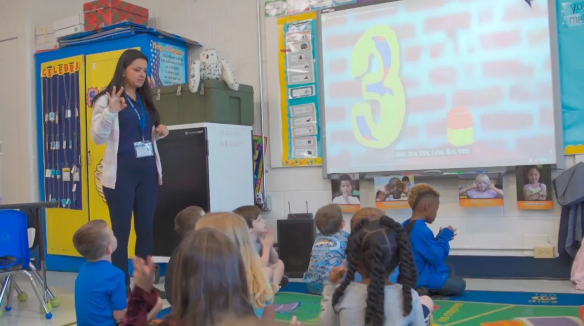 Female teacher smiles while holding both the Colombian and U.S. flags in front of a globe-themed backdrop. The photo reflects cross-cultural exchange, teaching in the United States, and professional development for educators abroad.