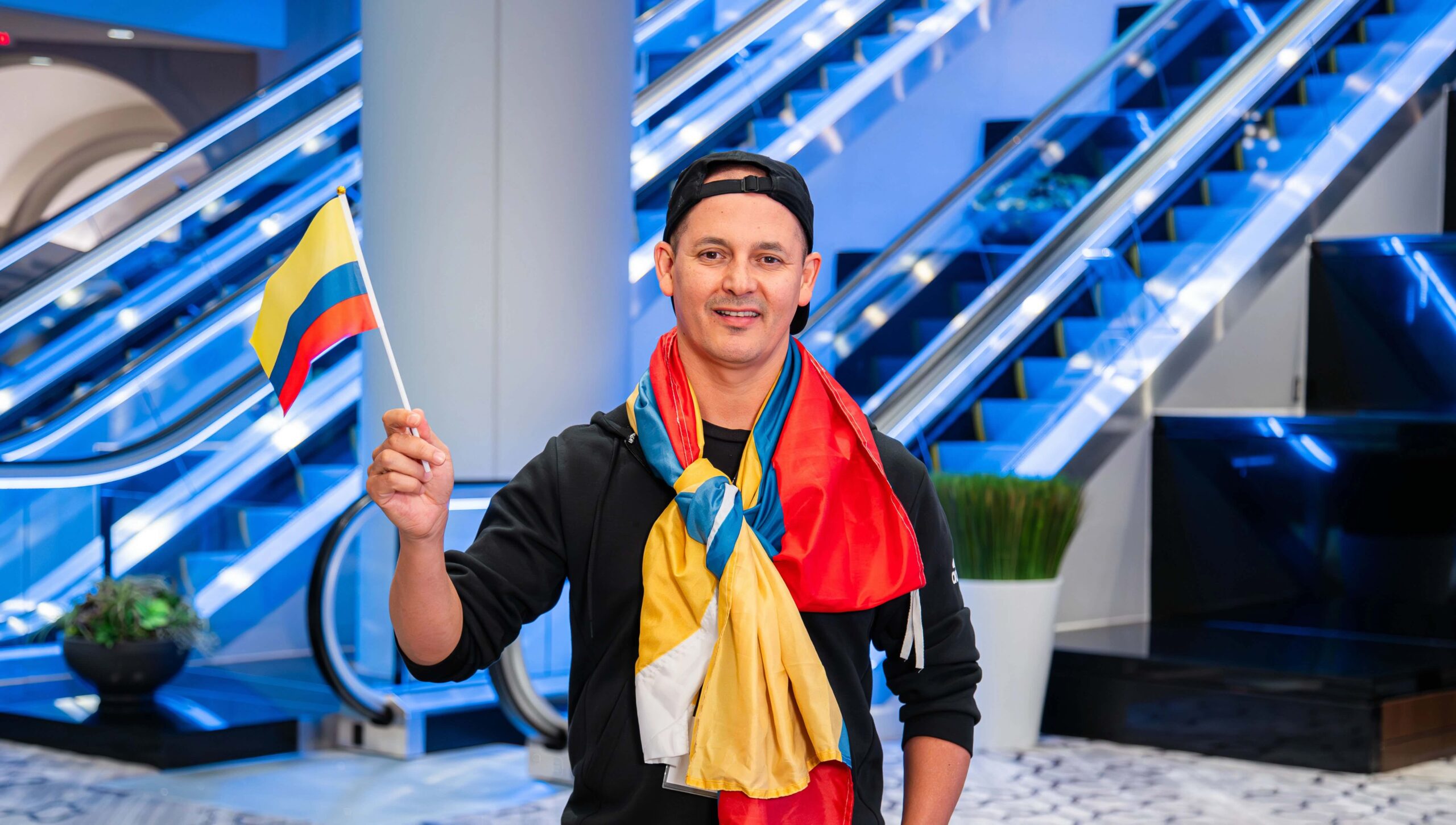 Smiling man wearing a backwards cap and draped in a Colombian flag proudly holds a small flag indoors, symbolizing teaching abroad and cultural exchange. Blue escalators and modern decor suggest a professional growth event tied to teaching in the United States.