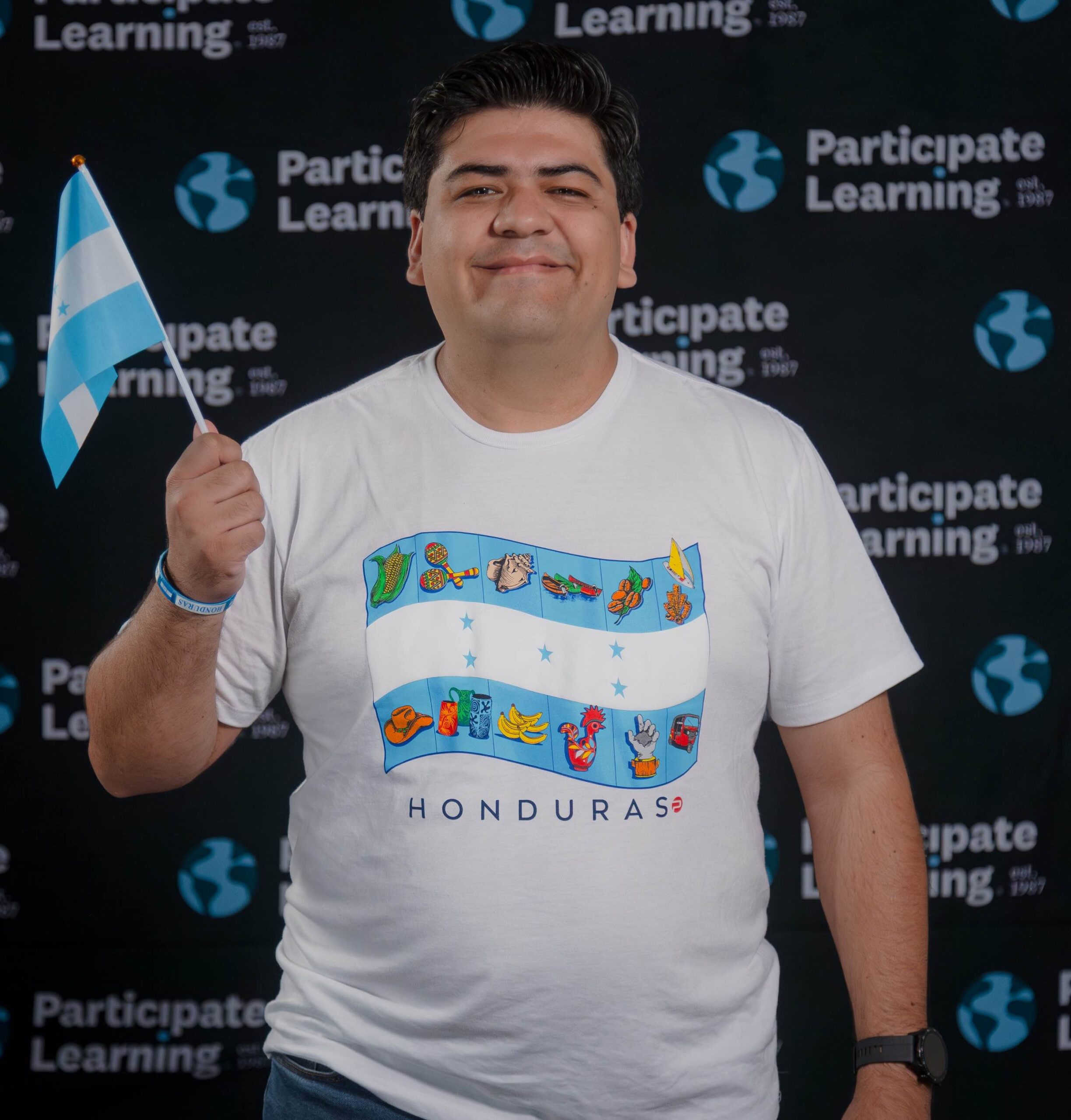 Smiling man holding a small Honduras flag stands in front of a Participate Learning backdrop, wearing a shirt with the Honduran flag and cultural icons. He is a new J-1 Cultural Exchange Teacher arriving to teach in the USA through with Participate Learning.