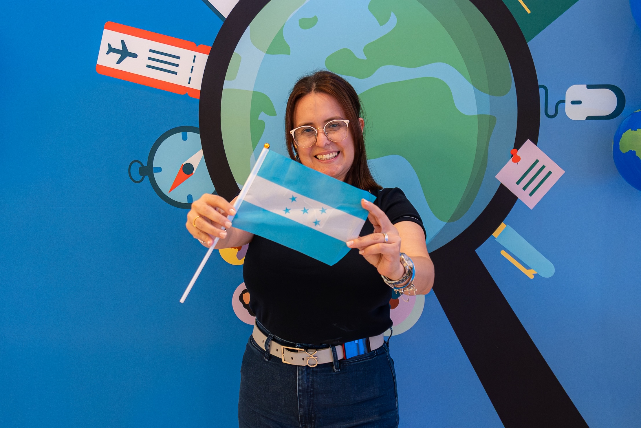 Smiling woman proudly holds a small Honduras flag in front of a colorful global education-themed backdrop, celebrating her arrival to teach in the USA with Participate Learning as part of a cultural exchange program.