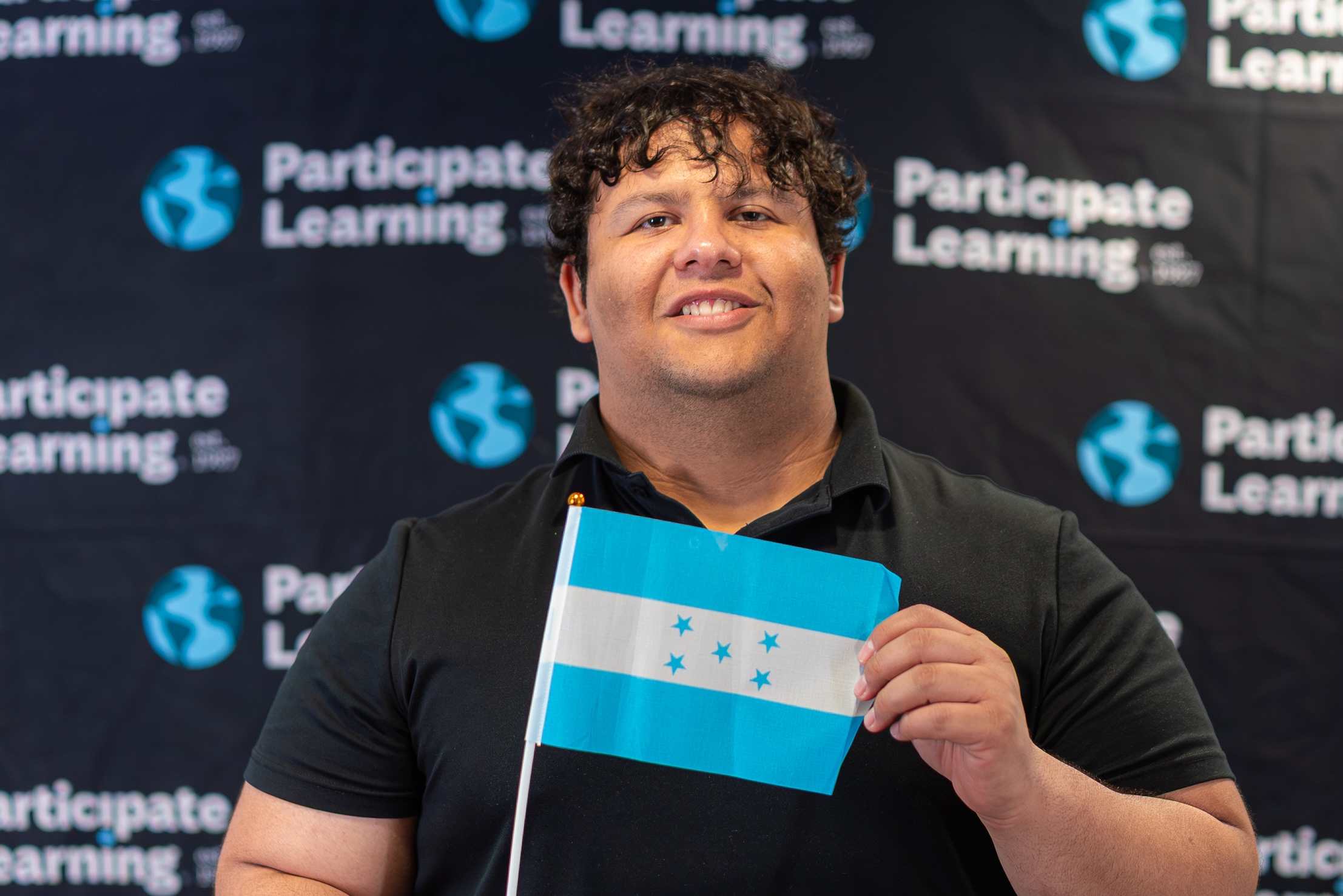 Man smiling and holding a small Honduras flag in front of a Participate Learning backdrop, marking his arrival to the U.S. to teach with Participate Learning as part of a cultural exchange teaching program.