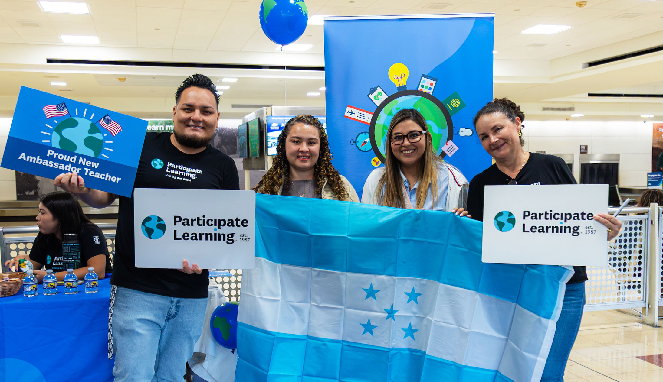 Four individuals from Honduras hold a national flag and Participate Learning signs as they arrive in the U.S. to teach with Participate Learning as Ambassador Teachers as part of a U.S. Department of State J-1 Cultural Exchange Teacher program.