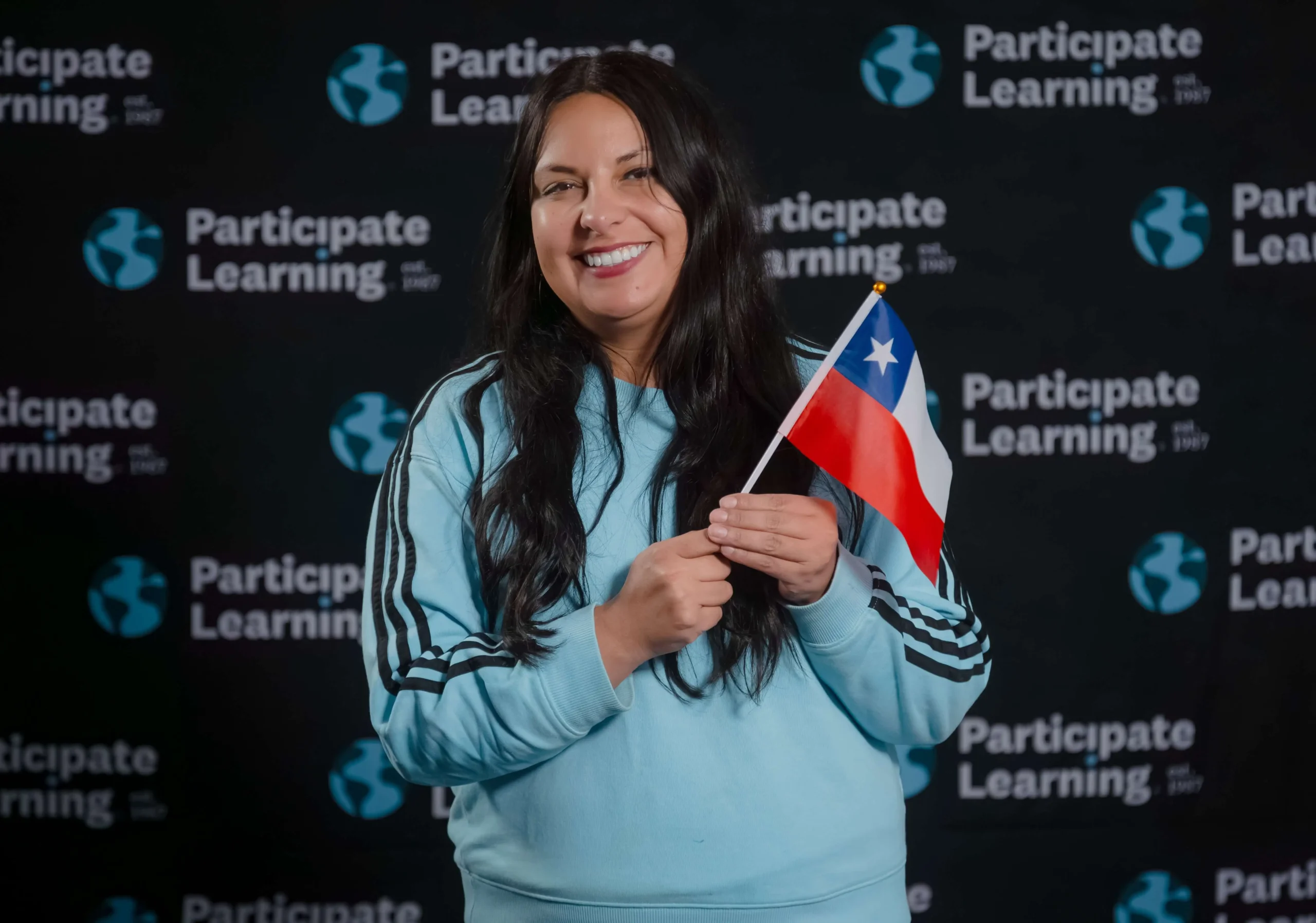 Female educator from Chile holding a small Chilean flag in front of a black Participate Learning branded backdrop. She is smiling confidently, representing her role as a teacher abroad.