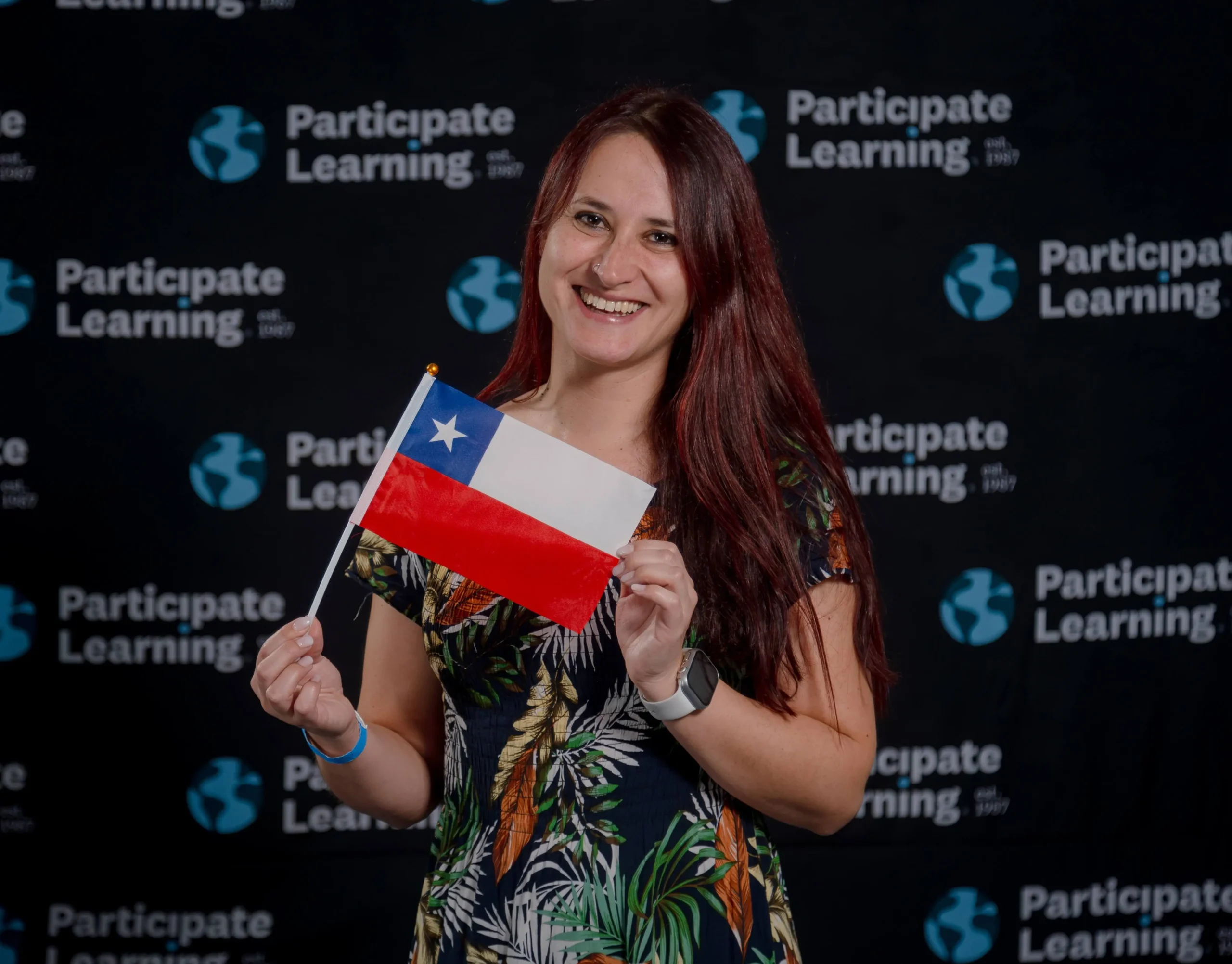 Chilean teacher with long red hair holding a Chilean flag in front of a Participate Learning banner, representing educators from Chile ready to teach in the USA through international exchange. She wears a floral print dress, suggesting a welcoming and festive atmosphere.