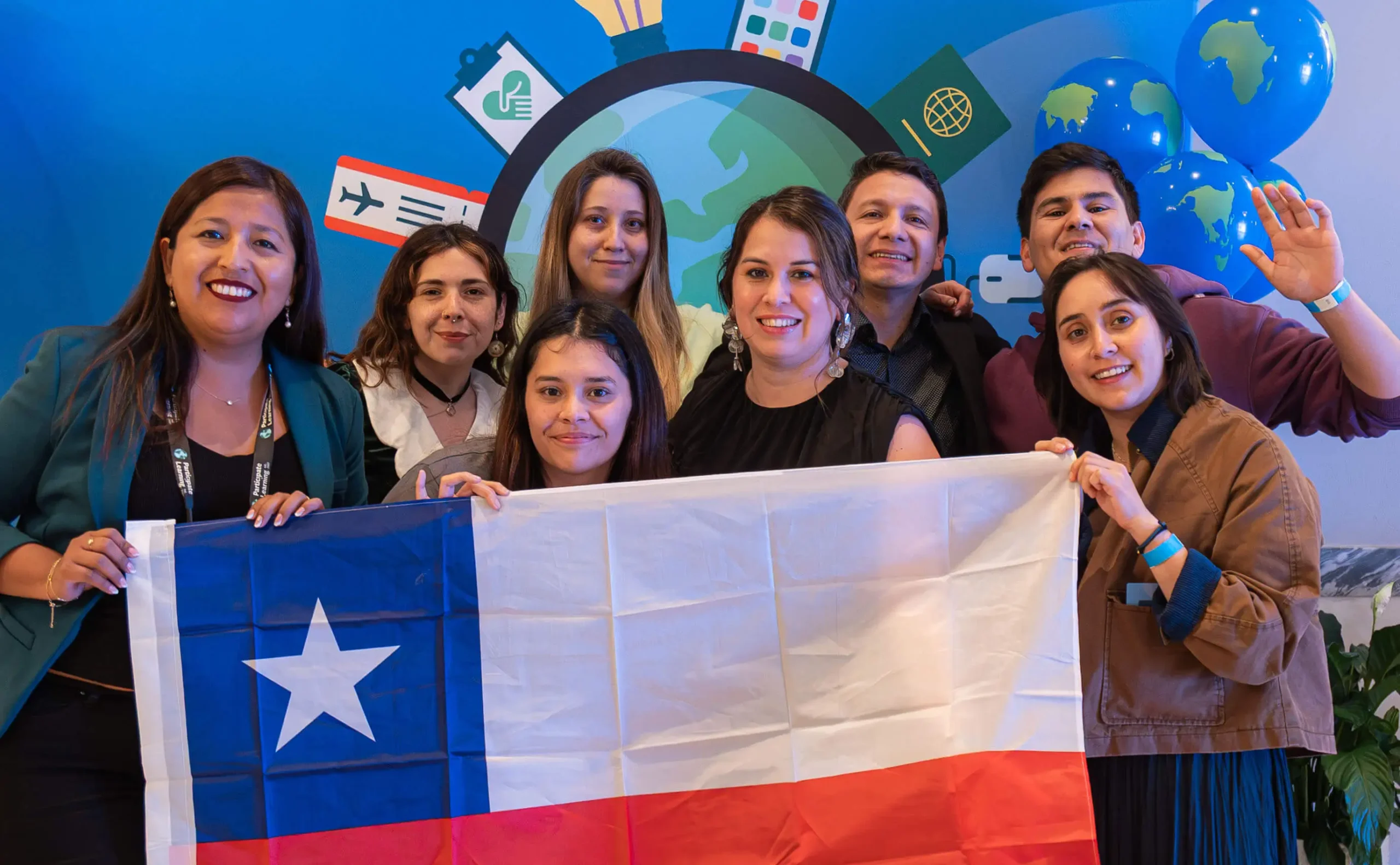 Group of eight smiling Chilean educators, including men and women, holding the Chilean flag during Ambassador Teacher Orientation. The colorful backdrop features travel and global education icons, representing Participate Learning's orientation.