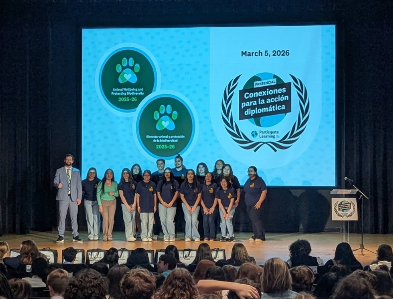 Middle school students stand on stage at UNC Greensboro during the Conexiones para la Acción Diplomática event as a presentation screen about animal wellbeing and biodiversity appears behind them.