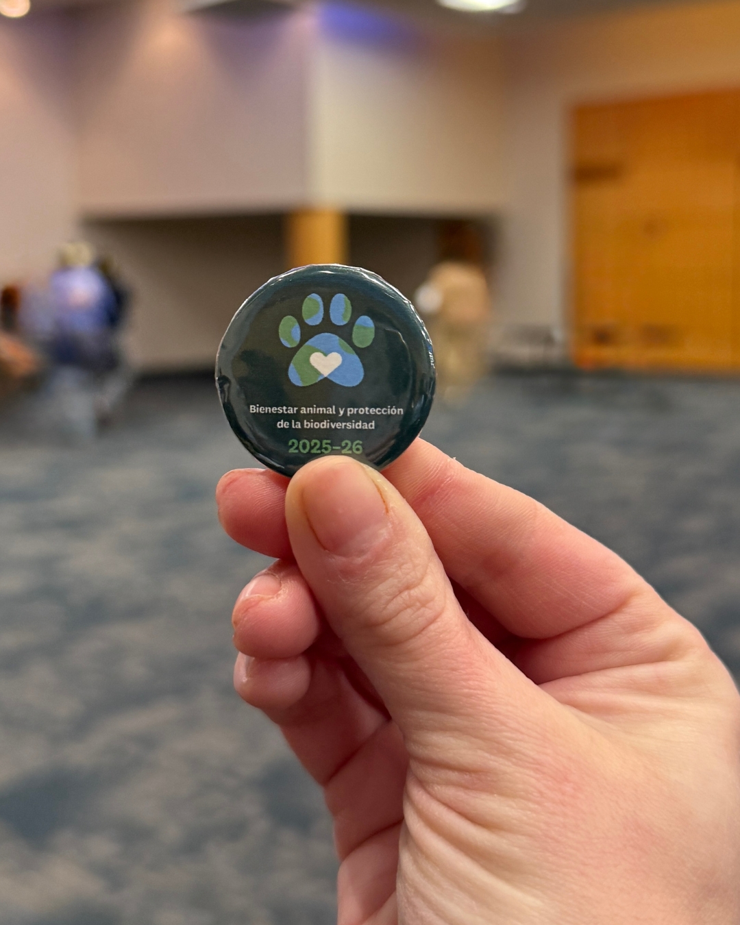 Hand holding a button that reads “Bienestar animal y protección de la biodiversidad 2025–26” with a paw print and heart symbol, representing the CAD event theme.
