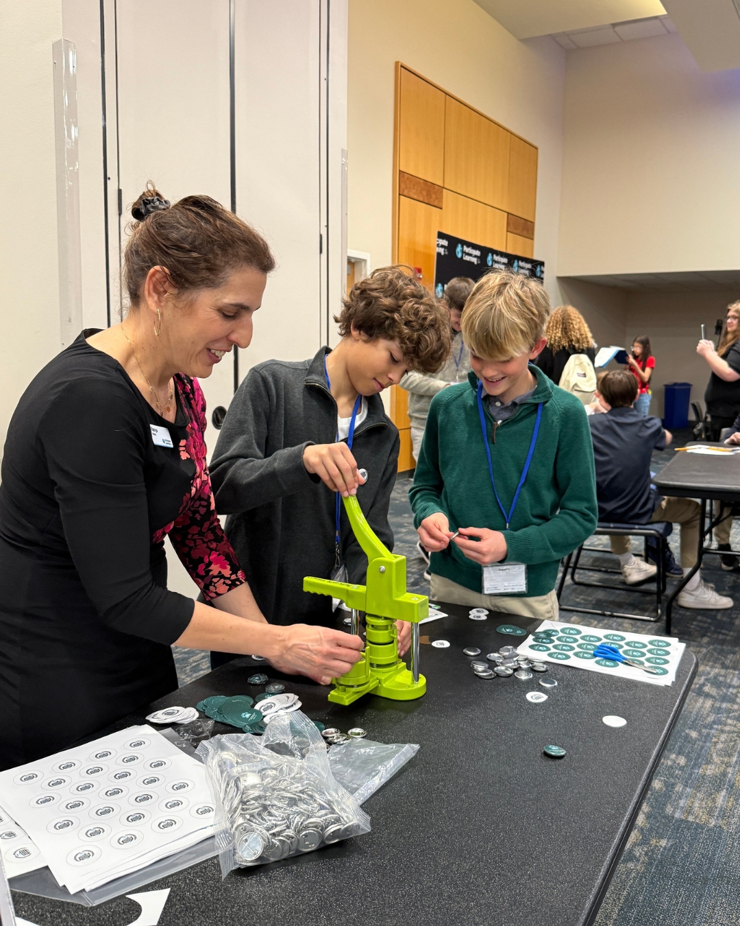 Two middle school students help a Participate Learning staff member use a button press to make event pins at a table covered with button parts during the CAD event.