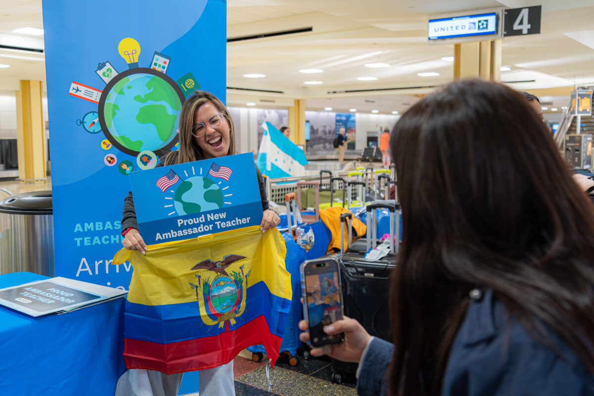 A teacher poses with the Flag of Ecuador and a "Proud New Ambassador Teacher" Participate Learning sign at the airport