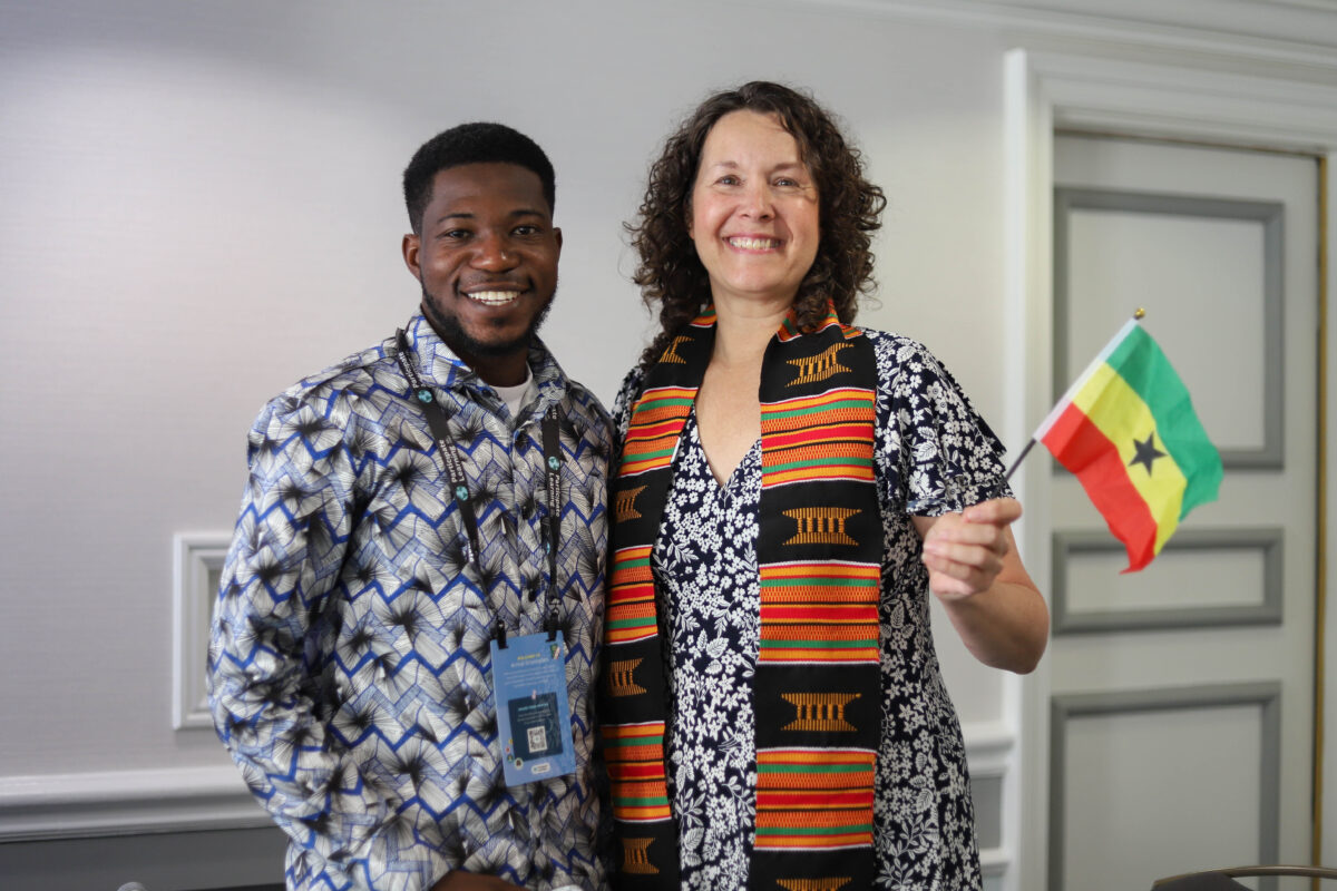 Two smiling adults pose together, one wearing a colorful woven stole and holding a Ghanaian flag.